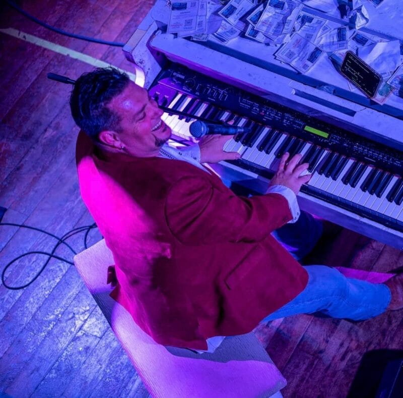 1 Grand Party Pop dueling piano entertainer in a red jacket playing a single piano covered in tip money during a lively crowd show.